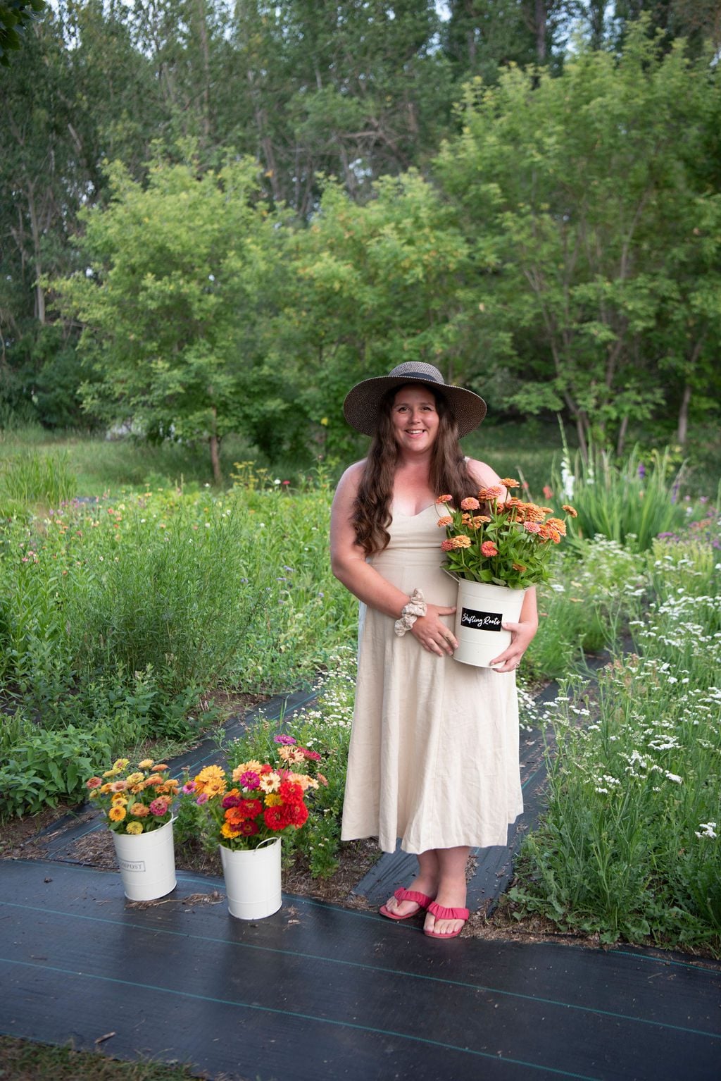 A woman in a beige dress and wide-brimmed hat stands in a garden holding a pot of orange zinnias. Two white buckets with flowers are on the ground beside her. Trees and greenery are in the background.