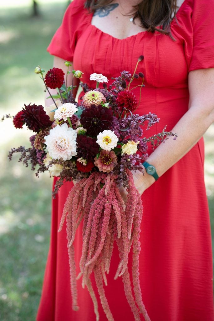 Woman in a red dress holds a bouquet of red, white, and pink flowers with long, trailing pink amaranthus, standing outdoors on grass.